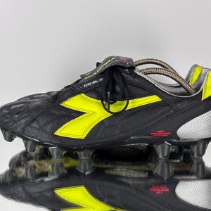 Op de afbeelding: Zwarte en gele voetbalschoen met een leren bovenwerk. De schoen heeft een geel chevron-ontwerp aan de zijkant en een zilveren hiel. De zool heeft meerdere noppen. De tong draagt de merknaam.