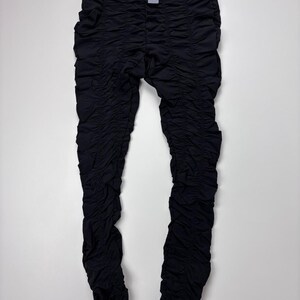 May include: Black leggings featuring a high-waisted design with a wide, smooth waistband and a small metallic logo tag. The entire length of the legs is covered in dense, horizontal ruching detail, creating a textured, gathered appearance.