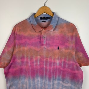 Puede incluir: Una camiseta polo de tie-dye en tonos rosa, naranja y azul. La camiseta tiene el logotipo de Polo Ralph Lauren en el pecho izquierdo.