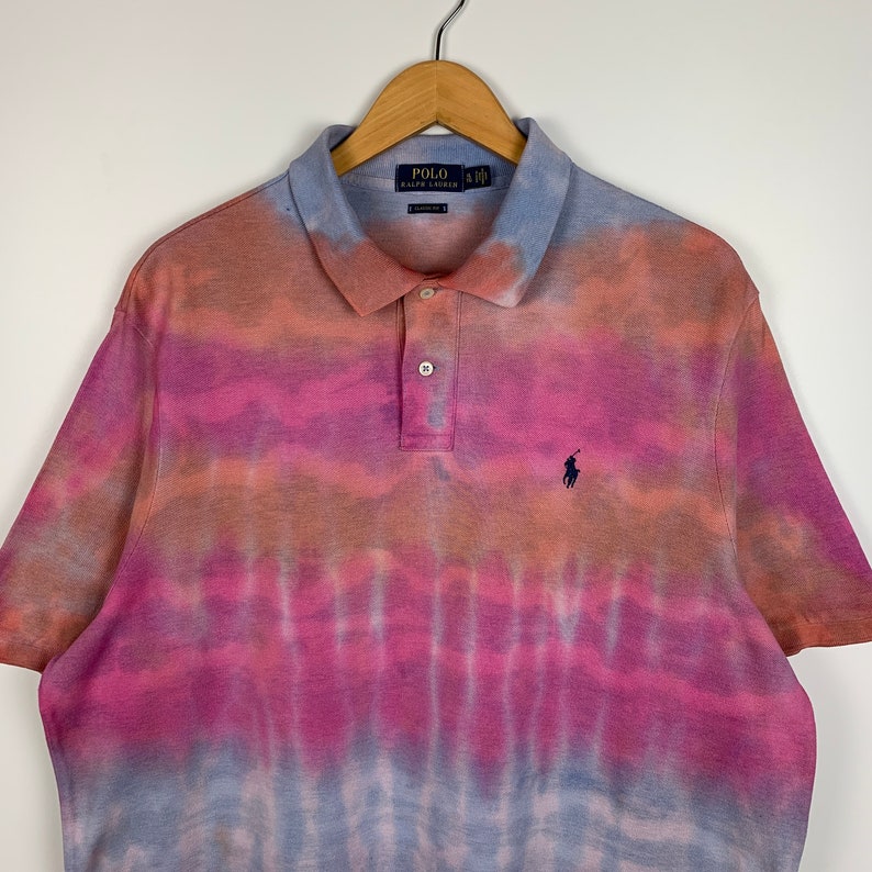 Puede incluir: Una camiseta polo de tie-dye en tonos rosa, naranja y azul. La camiseta tiene el logotipo de Polo Ralph Lauren en el pecho izquierdo.