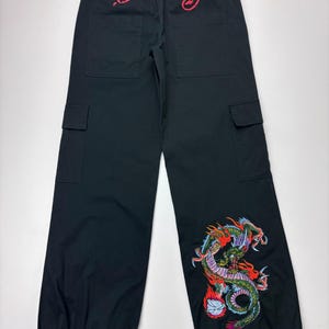 May include: Black cargo pants with a drawstring waist and ankle ties. The back features red embroidered text "Ed Hardy". A colorful dragon design is embroidered on the lower left leg. The pants have two front and two side pockets.