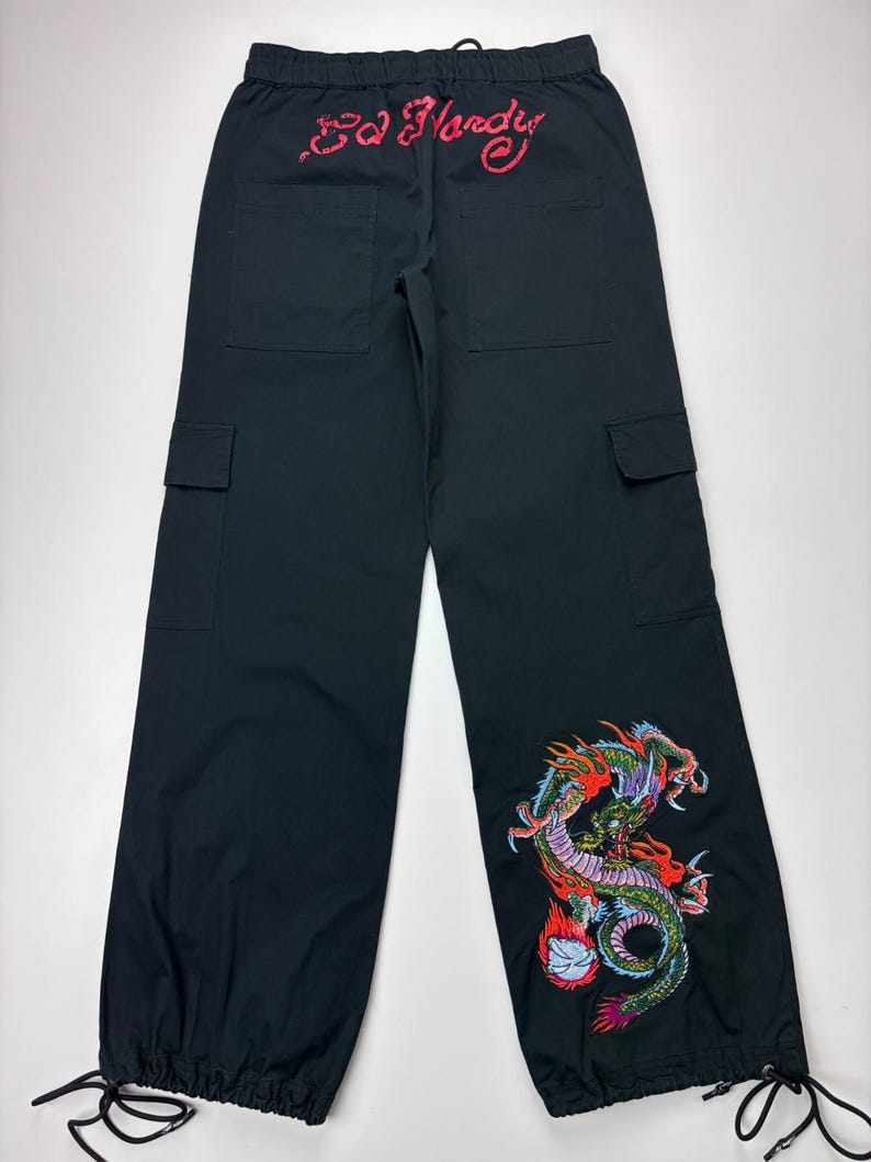 May include: Black cargo pants with a drawstring waist and ankle ties. The back features red embroidered text "Ed Hardy". A colorful dragon design is embroidered on the lower left leg. The pants have two front and two side pockets.