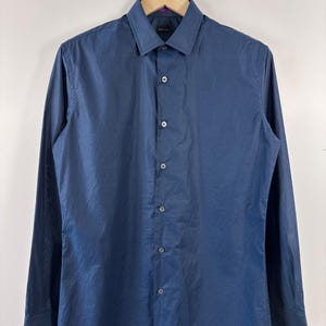 May include: A long-sleeved button-down shirt in a solid dark blue color. The shirt features a classic collar, a front button placket with matching dark blue buttons, and barrel cuffs. The fabric has a smooth, slightly sheen finish throughout.