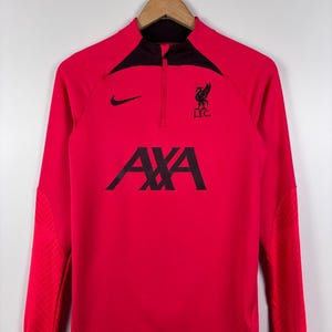 May include: A red long-sleeve athletic shirt with a black collar and a quarter-zip closure. The shirt features a black Nike swoosh, the Liverpool FC emblem, and the AXA logo. The shirt is hanging on a wooden hanger.