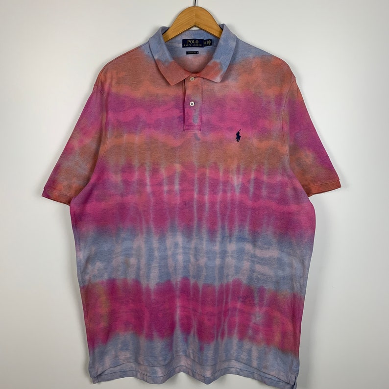 Puede incluir: Una camiseta polo de tie-dye en tonos de rosa, naranja y azul. La camiseta tiene un peque&ntilde;o logotipo bordado de jugador de polo en el pecho izquierdo.