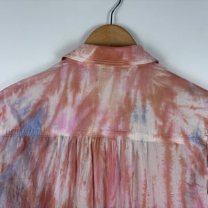 Pode incluir: Vista traseira de uma camisa abotoada com um padr&atilde;o tie-dye em tons de rosa, p&ecirc;ssego e azul. A camisa est&aacute; pendurada em um cabide de madeira contra um fundo branco. A gola e a pala das costas s&atilde;o vis&iacute;veis.