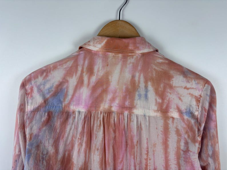 Pode incluir: Vista traseira de uma camisa abotoada com um padr&atilde;o tie-dye em tons de rosa, p&ecirc;ssego e azul. A camisa est&aacute; pendurada em um cabide de madeira contra um fundo branco. A gola e a pala das costas s&atilde;o vis&iacute;veis.