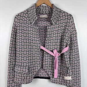 May include: A patterned cardigan in pink, black, and white, with a matching pink belt tied at the waist. The open-front sweater hangs on a wooden hanger. The cardigan has long sleeves and two pockets. The background is white.