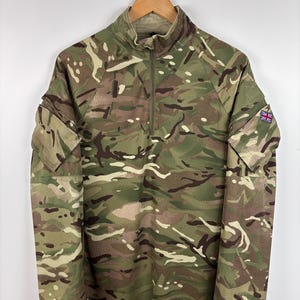 May include: A long-sleeved camouflage shirt with a half-zip collar. The shirt is patterned with green, brown, and tan camouflage. A small Union Jack patch is on the upper sleeve. The shirt is hanging on a wooden hanger.