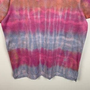 Puede incluir: Una camiseta tie-dye con un esquema de color azul, rosa y naranja. La camiseta tiene un corte holgado y un cuello redondo.