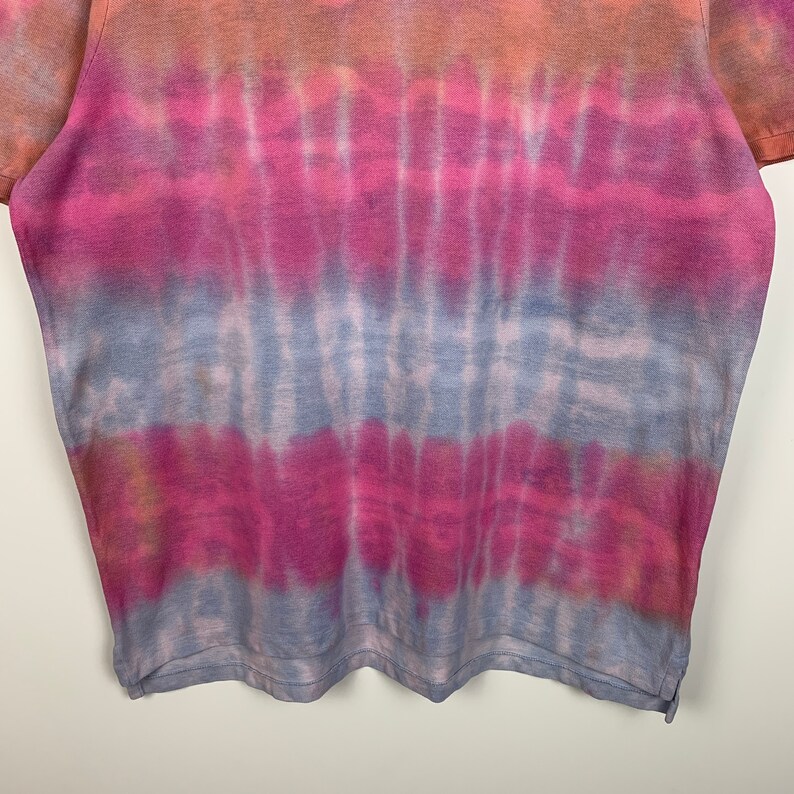 Puede incluir: Una camiseta tie-dye con un esquema de color azul, rosa y naranja. La camiseta tiene un corte holgado y un cuello redondo.