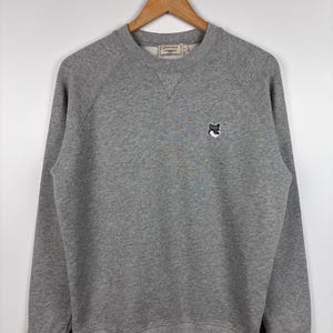 May include: A heather gray sweatshirt with a small fox logo on the chest. The crew neck sweatshirt is hanging on a wooden hanger. The sweatshirt has long sleeves and ribbed cuffs and hem.