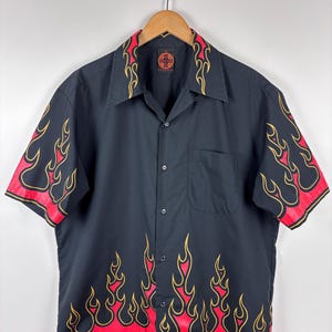 Könnte beinhalten: Schwarzes kurzärmeliges Hemd mit rotem und gelbem Flammendesign. Das Hemd hat einen klassischen Kragen und eine Tasche auf der linken Brust. Die Flammen sind gelb umrandet und haben eine rote Basis.