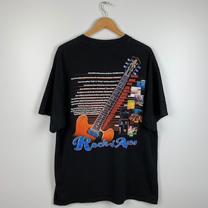 May include: Black t-shirt with a guitar graphic and the text "Rock of Ages" in blue. The design includes a timeline of rock music milestones. The shirt is hanging on a wooden hanger.