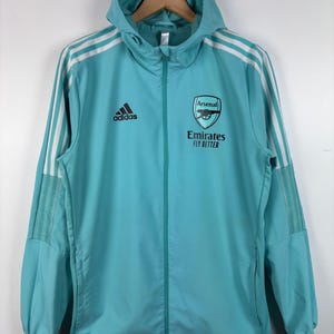 Könnte beinhalten: Türkisfarbene Kapuzenjacke mit weißen Streifen an den Ärmeln und dem Adidas-Logo auf der Brust. Das Arsenal-Teamlogo und "Emirates Fly Better" sind auf der rechten Brust aufgedruckt. Die Jacke hat einen durchgehenden Reißverschluss.