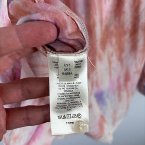 Pode incluir: Close-up de uma camisa tie-dye em tons de rosa, laranja e roxo. A etiqueta da camisa exibe instru&ccedil;&otilde;es de lavagem e informa&ccedil;&otilde;es de tamanho. A camisa tem um fecho de bot&atilde;o.
