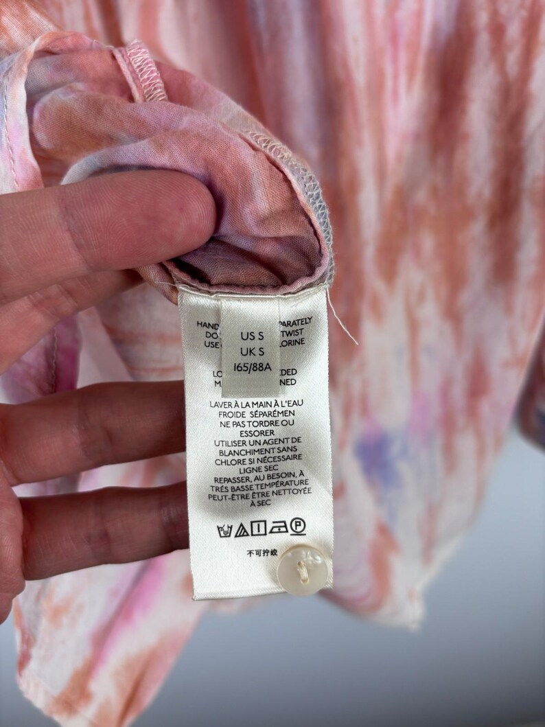 Pode incluir: Close-up de uma camisa tie-dye em tons de rosa, laranja e roxo. A etiqueta da camisa exibe instru&ccedil;&otilde;es de lavagem e informa&ccedil;&otilde;es de tamanho. A camisa tem um fecho de bot&atilde;o.