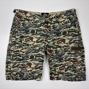 Pode incluir: Calças cargo camufladas com padrão marrom, verde e bege. As calças têm um fecho de correr e botão, e dois grandes bolsos cargo de cada lado.