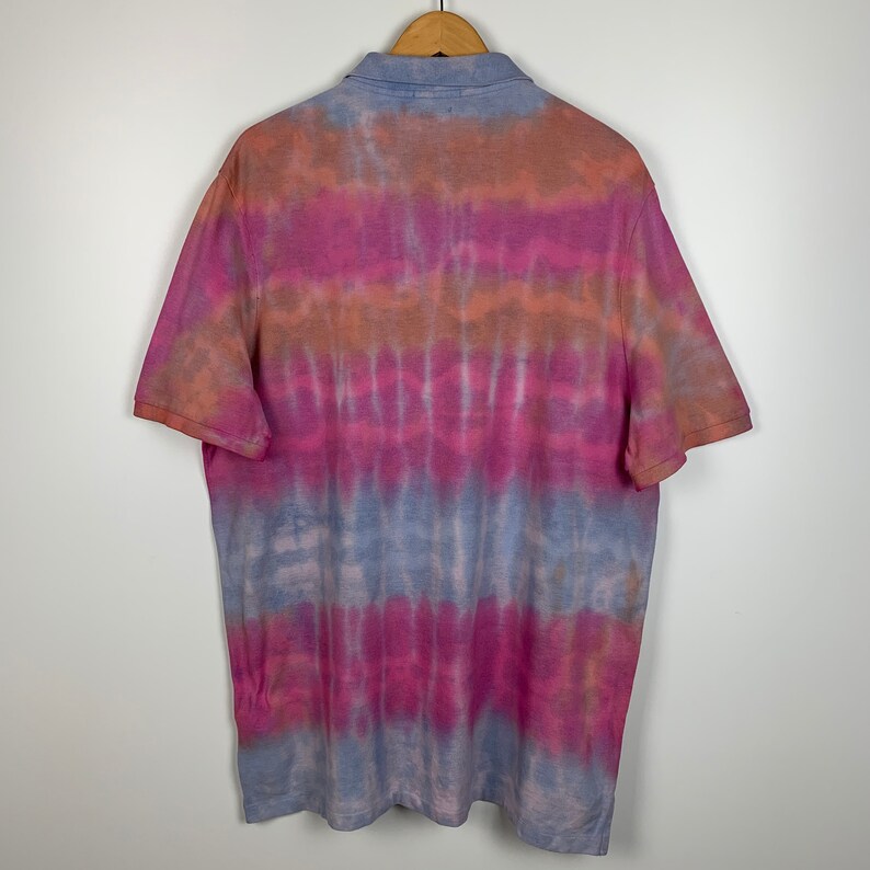 Puede incluir: Una camisa de manga corta con cuello y un patr&oacute;n de tie-dye en tonos de rosa, naranja y azul.