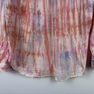 Pode incluir: Uma camisa de manga comprida com um padr&atilde;o tie-dye em tons de rosa, laranja e roxo. A camisa tem uma frente abotoada e punhos. O tecido parece leve e fluido, com um brilho subtil.