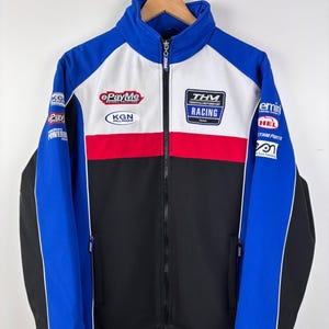 May include: A blue, white, red, and black zip-up racing jacket featuring a high collar and multiple sponsor logos including ePayMe, KGN, and THM Racing. The jacket has long sleeves, a full-length front zipper, and side pockets on a smooth fabric.