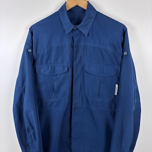 May include: A navy blue long-sleeve shirt with two chest pockets and buttoned cuffs. The shirt has a collared neckline and is hanging on a wooden hanger. A small white tag is visible on the right side of the shirt.