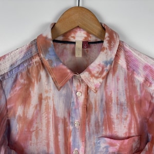 Pode incluir: Camisa abotoada com um padr&atilde;o tie-dye em tons de rosa, laranja e azul. A camisa tem gola, bolso no peito e mangas compridas. Est&aacute; pendurada num cabide de madeira.