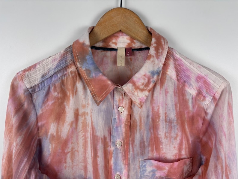 Pode incluir: Camisa abotoada com um padr&atilde;o tie-dye em tons de rosa, laranja e azul. A camisa tem gola, bolso no peito e mangas compridas. Est&aacute; pendurada num cabide de madeira.
