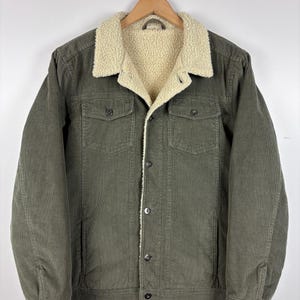 May include: Olive green corduroy jacket with a cream-colored sherpa lining and collar. The jacket has two chest pockets, two side pockets, and a button-up front. The jacket is hanging on a wooden hanger.