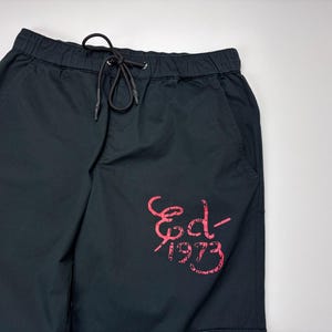 May include: Black cargo shorts with an elastic waistband and drawstring. The shorts feature a pink graphic on the leg that reads "Ed-1973". The shorts have pockets and are made of a durable fabric.