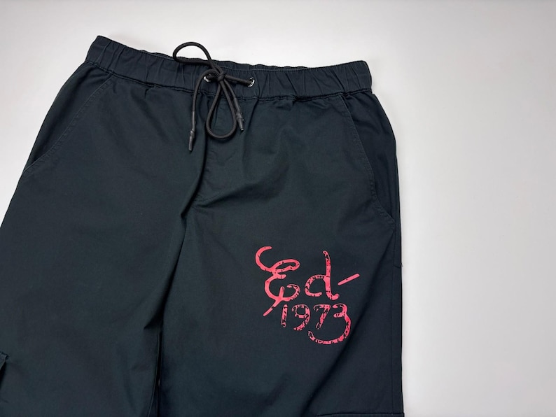 May include: Black cargo shorts with an elastic waistband and drawstring. The shorts feature a pink graphic on the leg that reads "Ed-1973". The shorts have pockets and are made of a durable fabric.