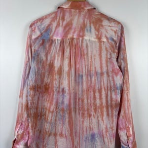 Pode incluir: Camisa de manga comprida com padr&atilde;o tie-dye em tons de rosa, laranja e roxo. A camisa est&aacute; pendurada num cabide de madeira sobre um fundo branco. Possui gola cl&aacute;ssica e punhos abotoados.