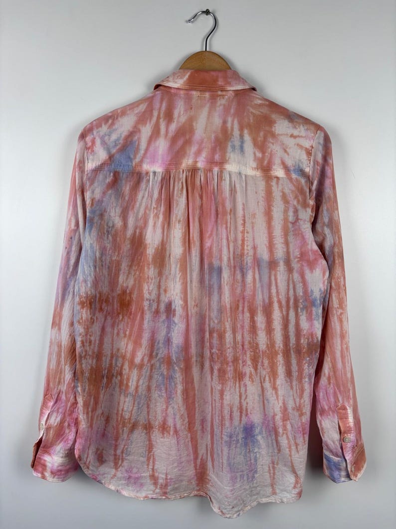 Pode incluir: Camisa de manga comprida com padr&atilde;o tie-dye em tons de rosa, laranja e roxo. A camisa est&aacute; pendurada num cabide de madeira sobre um fundo branco. Possui gola cl&aacute;ssica e punhos abotoados.