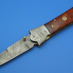 以下が含まれることがあります： 模様入りの鋼の刃と木製の柄が付いた折りたたみナイフ。 柄は濃い茶色で、真鍮のアクセントと装飾的な円形インレイが付いています。 刃は開いており、独特の波のような模様を示しています。