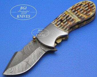 May include: A folding pocket knife with a Damascus steel blade and a brown and white bone handle. The knife has a bottle opener and the words "BGI Handcrafted USA KNIVES" are engraved on the handle.
