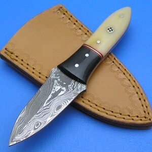 May include: A small, black-handled knife with a damascus steel blade and a bone handle. The knife is in a brown leather sheath.