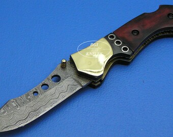 May include: A folding knife with a patterned blade and a wooden handle. The handle is a dark red and black color, with brass accents. The blade has a unique wave-like pattern. The knife is on a blue background.