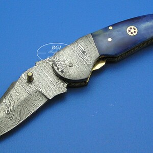May include: A folding pocket knife with a Damascus steel blade and a blue and white bone handle. The handle has a gold colored accent and a red and gold emblem.