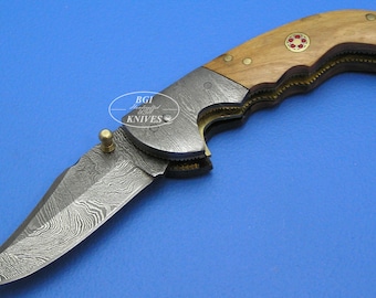 May include: A folding pocket knife with a wood handle and a Damascus steel blade. The handle has a brass inlay and a red and gold emblem. The blade is partially open and shows the intricate pattern of the Damascus steel.
