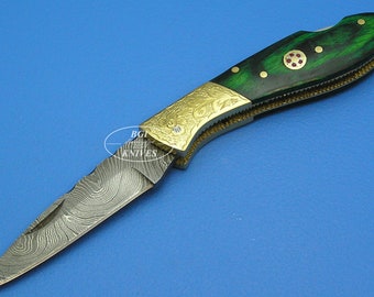May include: A folding pocket knife with a green wood handle and a damascus steel blade. The handle has gold accents and a red jewel inlay.