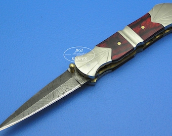 May include: A red and silver folding knife with a damascus steel blade. The handle is made of wood and has a decorative design. The knife is closed and has a safety lock.