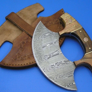 Puede incluir: Un cuchillo curvo de color plateado con mango de madera y funda de cuero. El mango tiene un diseño decorativo con piedras preciosas rojas.