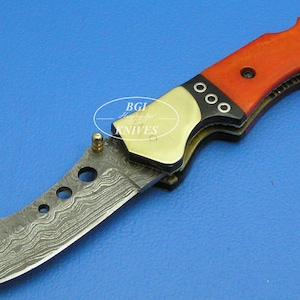 May include: A red and black folding knife with a Damascus steel blade. The handle is made of wood and has a brass accent.