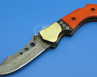May include: A red and black folding knife with a Damascus steel blade. The handle is made of wood and has a brass accent.