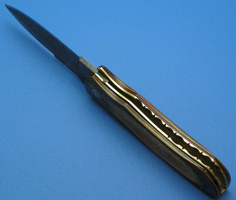 May include: A handcrafted folding knife with a dark blade and a handle featuring a multi-colored wood grain design. The handle is accented with brass and a decorative pattern.