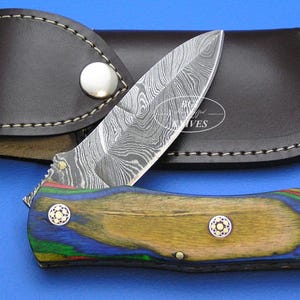 May include: A folding knife with a patterned blade and a wooden handle with blue, green, and yellow accents. The knife is paired with a brown leather sheath. The blade has a unique, swirling pattern. The handle has two decorative rivets.