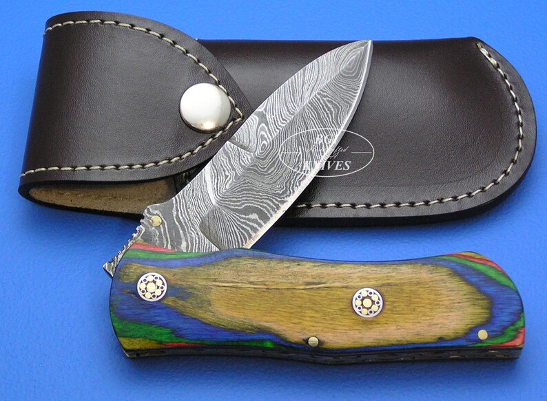 May include: A folding knife with a patterned blade and a wooden handle with blue, green, and yellow accents. The knife is paired with a brown leather sheath. The blade has a unique, swirling pattern. The handle has two decorative rivets.