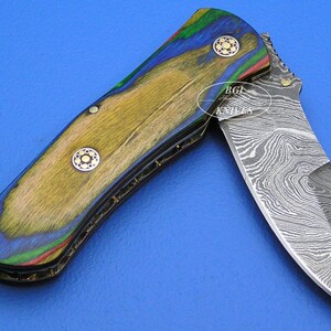May include: A partially open folding knife with a patterned steel blade and a handle made of wood with a colorful design. The handle features shades of blue, green, red, and yellow. The knife has decorative rivets.