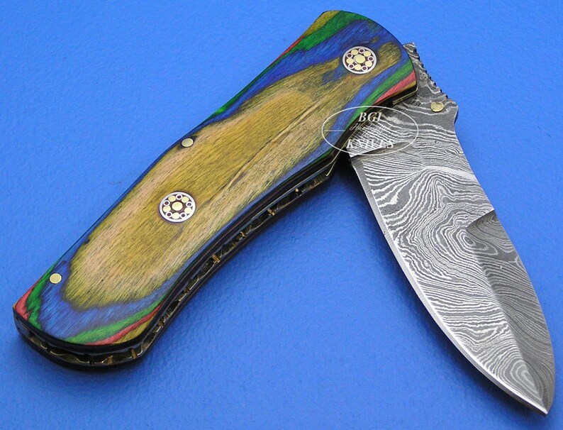 May include: A partially open folding knife with a patterned steel blade and a handle made of wood with a colorful design. The handle features shades of blue, green, red, and yellow. The knife has decorative rivets.