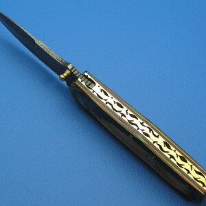 May include: A handcrafted folding knife with a dark blade and a handle featuring a gold-colored, leaf-like design. The handle has a dark wood appearance with gold accents. The knife is set against a blue background.